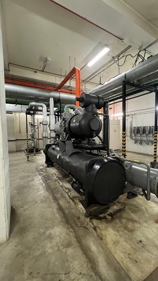 A water-cooled chiller in a plant room: the compressor sits above a long horizontal shell, with insulated pipework and an orange support frame behind.